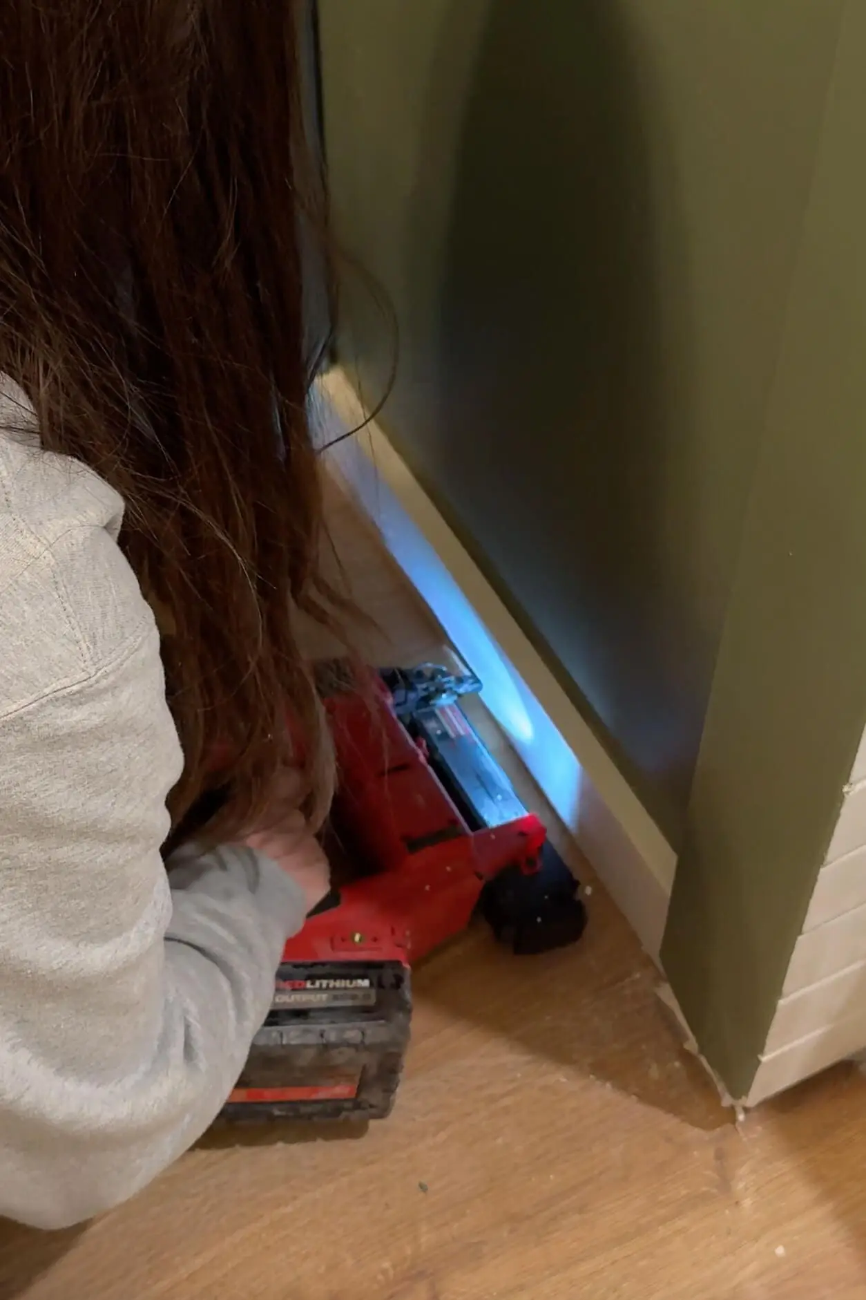 Close-up of a person using a red nail gun to secure baseboard trim on a green bathroom wall, capturing the precision and tools involved in DIY woodworking.